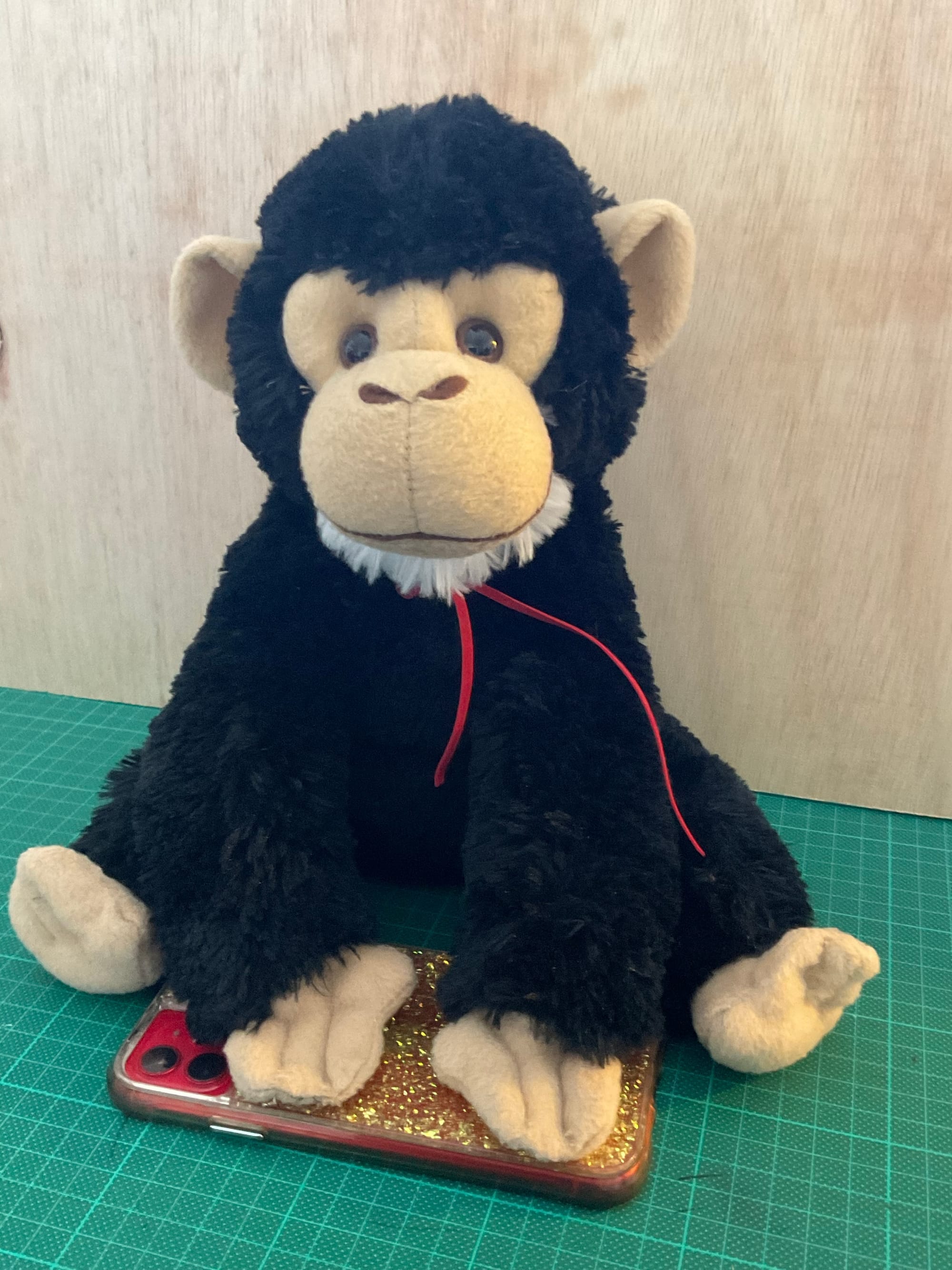 A stuffed howler monkey sits on a green cutting mat with its paws resting on a red smart phone in a glittery case. 